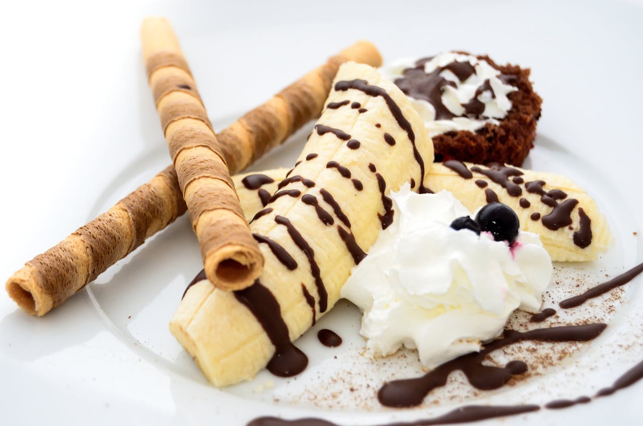 creative-02 Close-up of a banana dessert with chocolate drizzle, whipped cream, and wafer sticks.