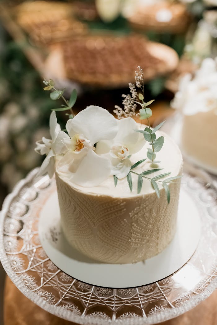 brand-03 A beautifully decorated wedding cake adorned with white flowers and leaves on a glass stand.